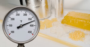 Close up of a temperature gauge in front of a table with different types of hot melt adhesives on it.